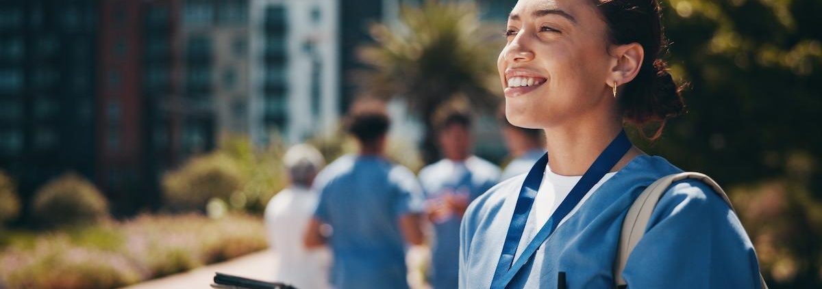 A happy, smiling female healthcare professional in blue scrubs is standing outdoors on a sunny day. She is holding a clipboard or tablet and looking up and to the right. Her hair is tied back, and she is wearing small hoop earrings. In the blurred background, other medical personnel are walking near modern, multi-story buildings and trees.