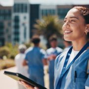 A happy, smiling female healthcare professional in blue scrubs is standing outdoors on a sunny day. She is holding a clipboard or tablet and looking up and to the right. Her hair is tied back, and she is wearing small hoop earrings. In the blurred background, other medical personnel are walking near modern, multi-story buildings and trees.