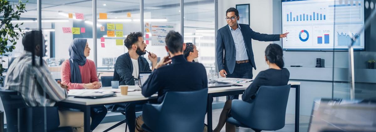 An image of a diverse group of business professionals sitting around a table in a bright, modern office for a meeting. A male presenter in a suit is standing and pointing at a large wall-mounted screen displaying charts and data visualizations. Five other colleagues are seated, actively listening and engaging, including a woman wearing a hijab. The office features glass walls and sticky notes with various ideas.