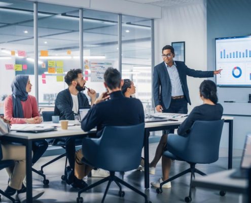 An image of a diverse group of business professionals sitting around a table in a bright, modern office for a meeting. A male presenter in a suit is standing and pointing at a large wall-mounted screen displaying charts and data visualizations. Five other colleagues are seated, actively listening and engaging, including a woman wearing a hijab. The office features glass walls and sticky notes with various ideas.