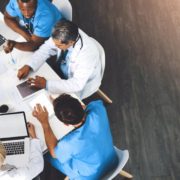 An overhead view shows a group of five medical professionals sitting around a white table during a meeting. They are collaborating and looking at documents, laptops, and tablets spread across the table. Two individuals wear blue scrubs, and three wear white lab coats or professional attire. The floor is dark wood, and a bright light source comes from the upper right corner.
