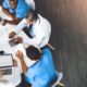 An overhead view shows a group of five medical professionals sitting around a white table during a meeting. They are collaborating and looking at documents, laptops, and tablets spread across the table. Two individuals wear blue scrubs, and three wear white lab coats or professional attire. The floor is dark wood, and a bright light source comes from the upper right corner.