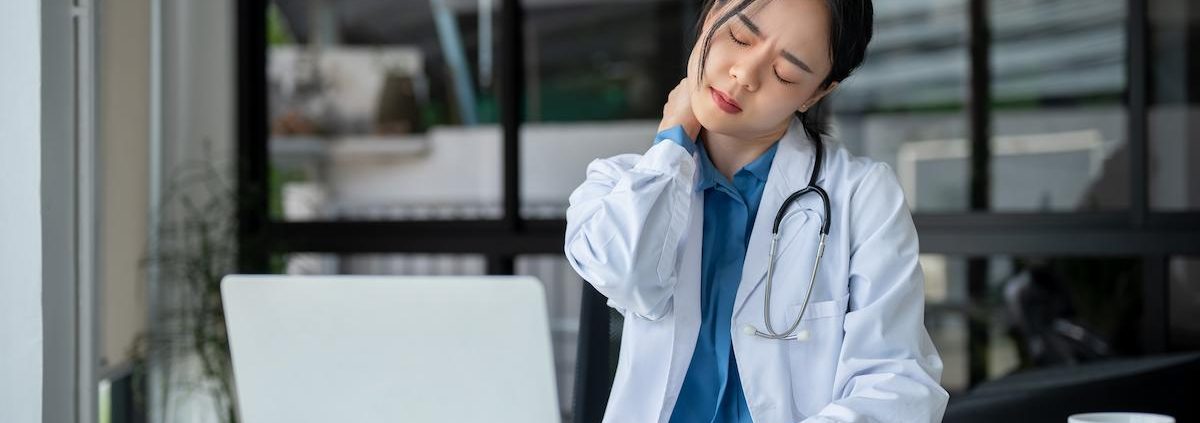 A female doctor or medical professional in a white lab coat and stethoscope is seated at a desk, showing signs of stress or neck pain. She is holding the back of her neck and grimacing, with her eyes closed. A laptop, clipboard with documents, and a coffee cup are on the desk in front of her. The setting appears to be an office with large windows in the background.
