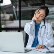 A female doctor or medical professional in a white lab coat and stethoscope is seated at a desk, showing signs of stress or neck pain. She is holding the back of her neck and grimacing, with her eyes closed. A laptop, clipboard with documents, and a coffee cup are on the desk in front of her. The setting appears to be an office with large windows in the background.