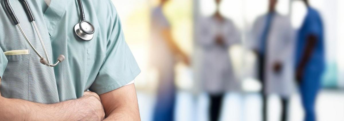 A cropped, close-up shot of a medical professional standing with their arms crossed. They are wearing light green scrubs and a stethoscope draped around their neck. The background is blurred, showing a group of four other indistinct medical staff members standing in a well-lit indoor area.